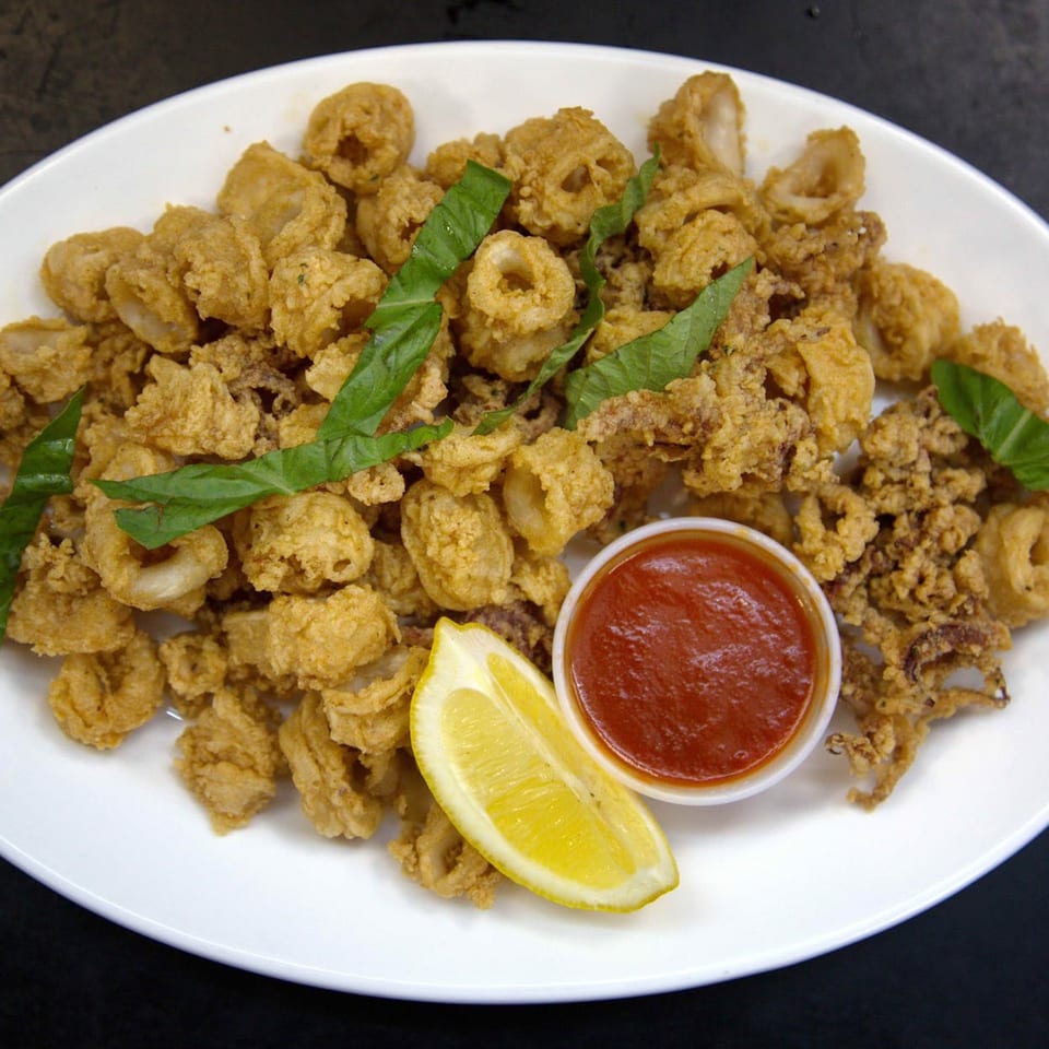 Crispy fried calamari with fresh basil, a lemon wedge, and marinara sauce on a white plate.