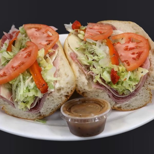 Two Italian subs with deli meat, cheese, lettuce, tomatoes, and peppers, with a side of dressing.
